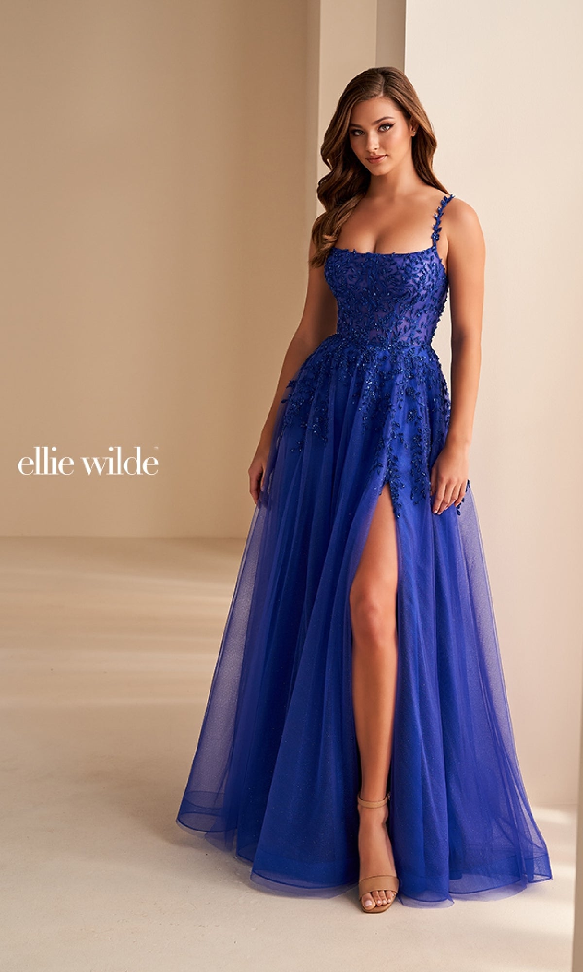 A woman stands indoors against a neutral background wearing the Long Prom Dress: Ellie Wilde EW36228, a royal blue, sleeveless, floor-length gown with a corset bodice, floral embellishments, and an elegant thigh-high slit.